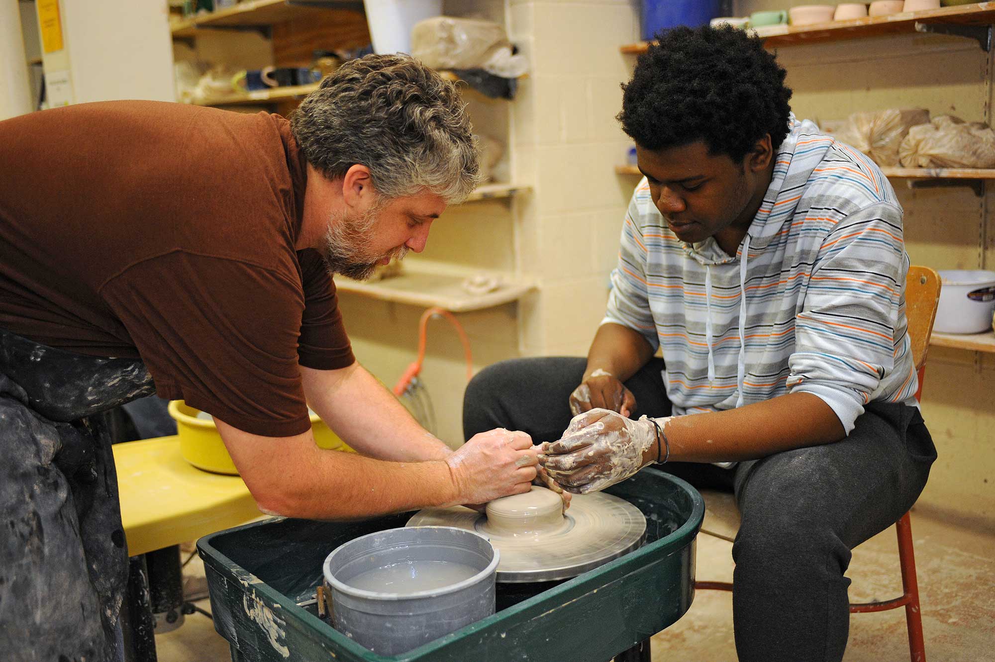 Professor helps student throw pot on potter's wheel.