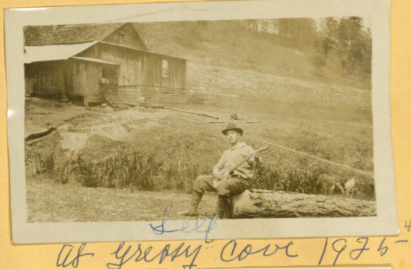 man sitting on log with banjo outside with house nearby