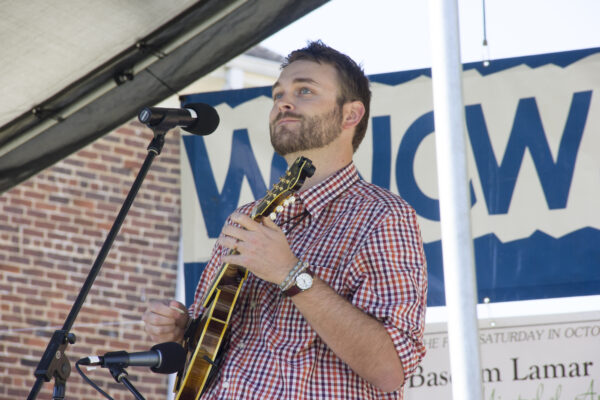 Young man in checkered shirt playing mandolin on stage.