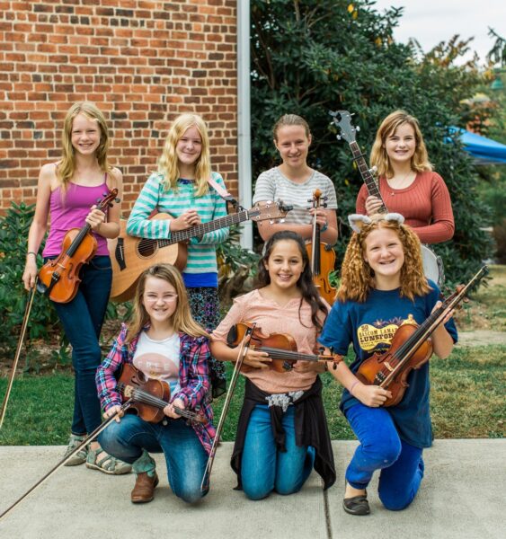 seven young musicians posing outside holding fiddles and guitars and one banjo.