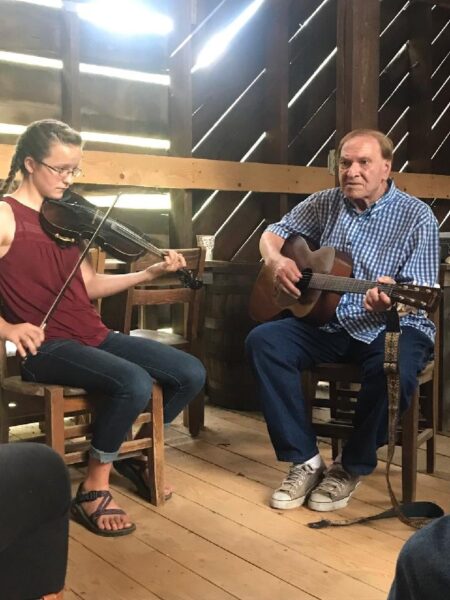 Young woman fiddling next to older man who is playing guitar. They are in a barn.