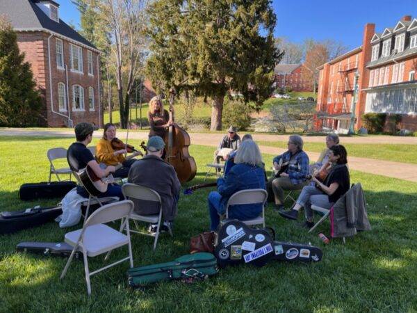 A group of fiddlers site in folding chairs in a circle in the shade with two buildings and a tree in the sunlit area behind them.