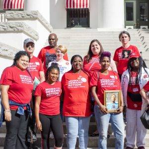 Ten people, most wearing red t-shirts, stand on the steps of the Capitol. One person is holding a framed photo of someone killed by gun violence.