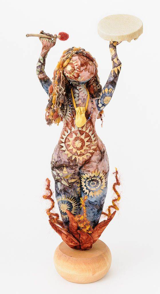 doll figure of a person with long hair holding a tambourine and a drum mallet; the figure has multi-colored sunburtsts adorning the torso, arms, and legs