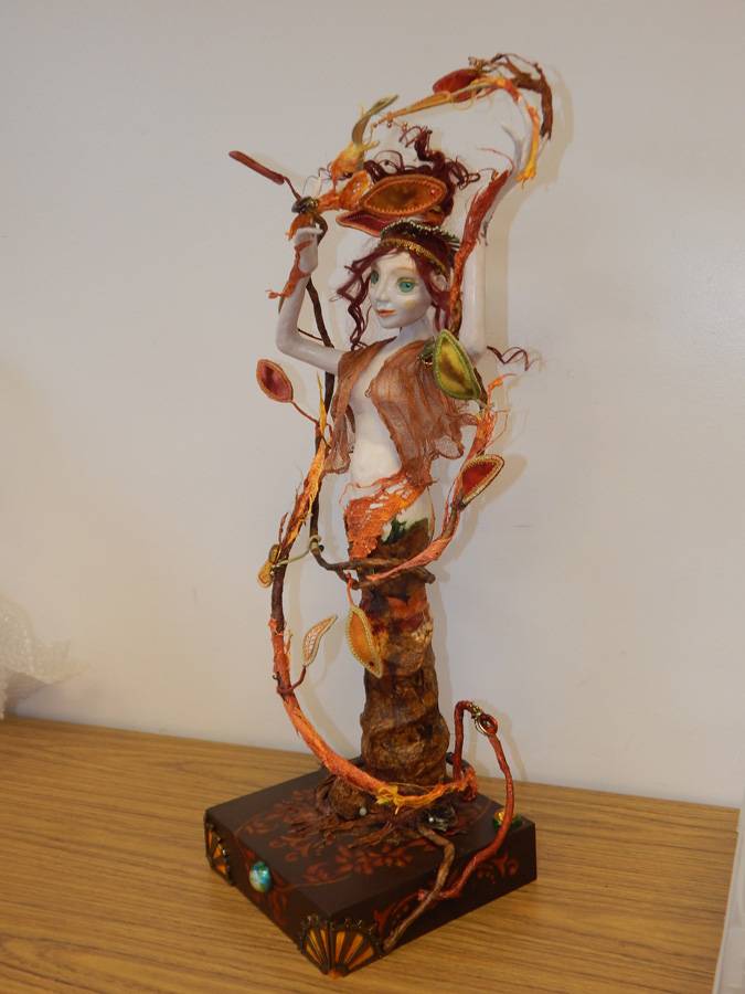 A doll in autumnal colors of orange and brown is affixed to a pedestal. The figure appears to grow from a tree stump with leaves and vines entwined.