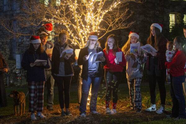 Carolers stand in front of a tree with white Christmas lights and sing.