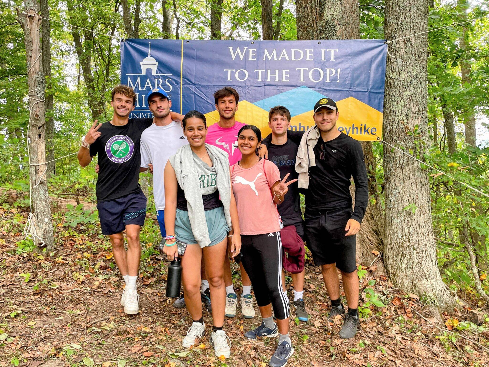 A group of seven students stands in front of the "We Made It To the Top" banner at the top of Bailey Mountain.