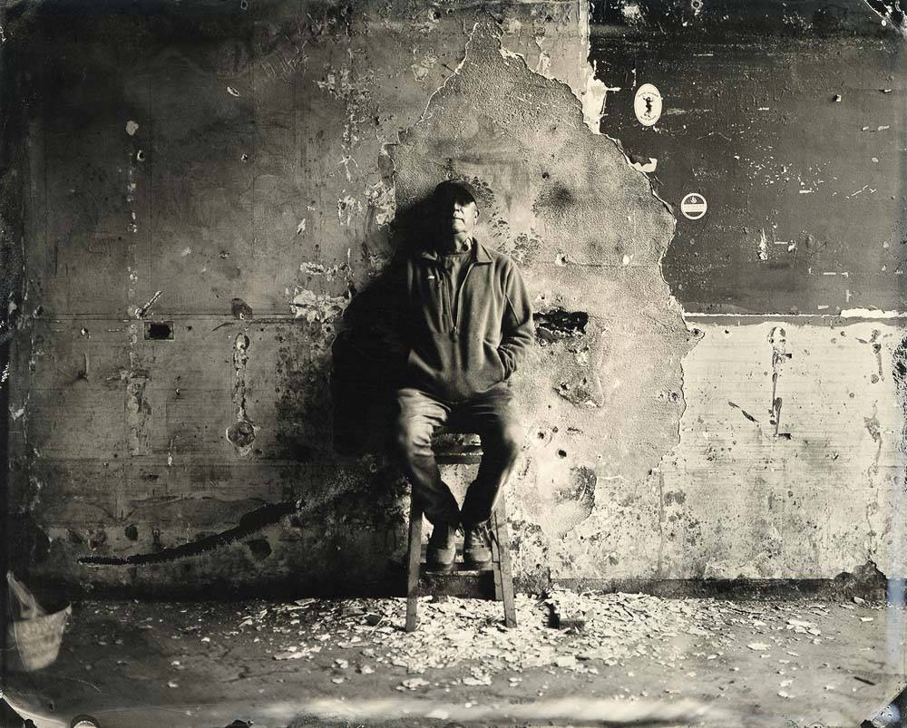 Tintype photograph of a man standing against a wall damaged by flood waters