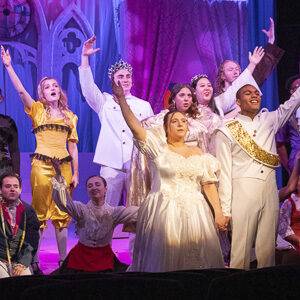 6 performers on stage dressed in costume for Cinderella. The females are wearing yellow and the man in front is dressed like a prince. They have one arm outstretched as they sing. 