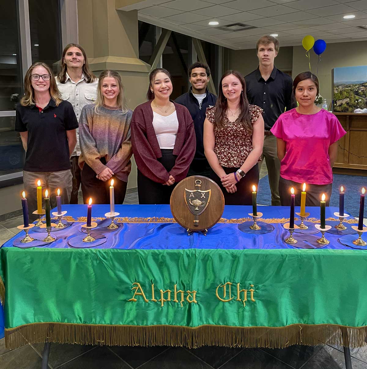 Alpha Chi inductees line up in two rows behind a table with candles, a plaque, and a green and blue Alpha Chi tablecloth.