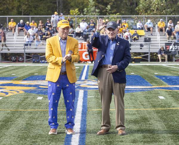 Wayne Higgins waves to the crowd during his presentation as 2025 Alumnus of the Year during halftime of the homecoming football game. Pres. Floyd stands with him.